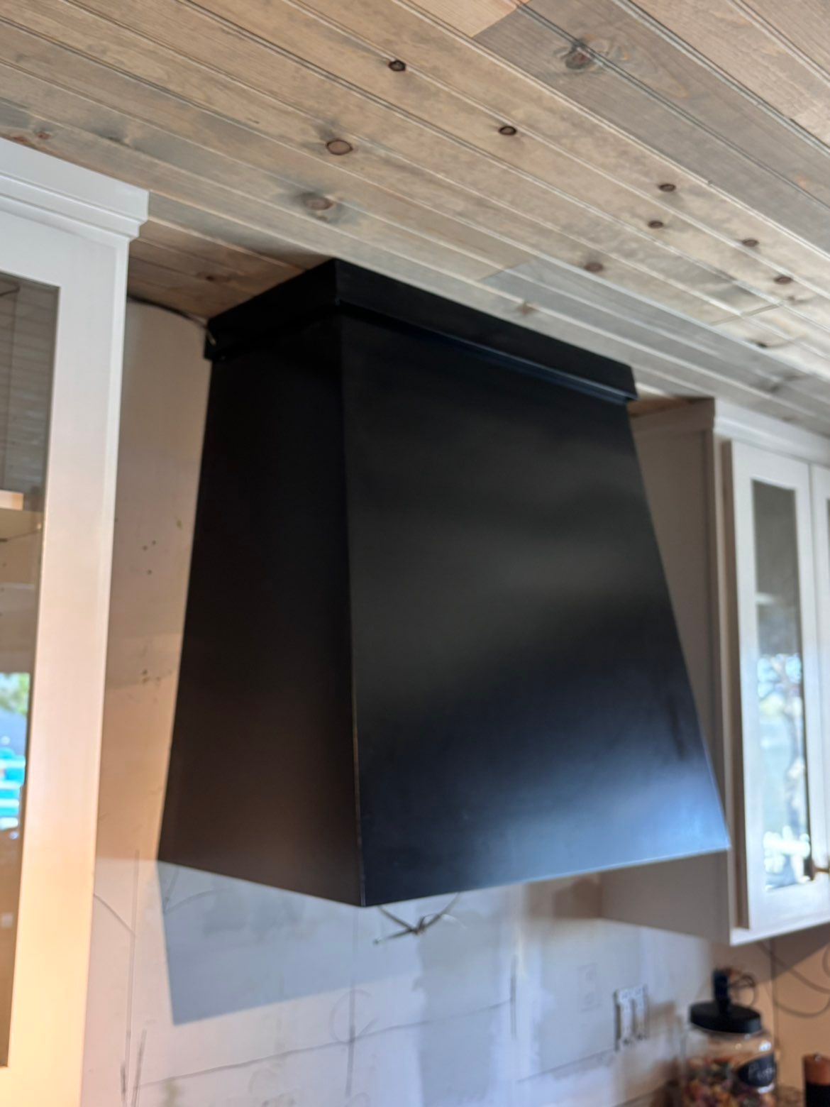Black range hood mounted on a kitchen wall beneath a wood-paneled ceiling.