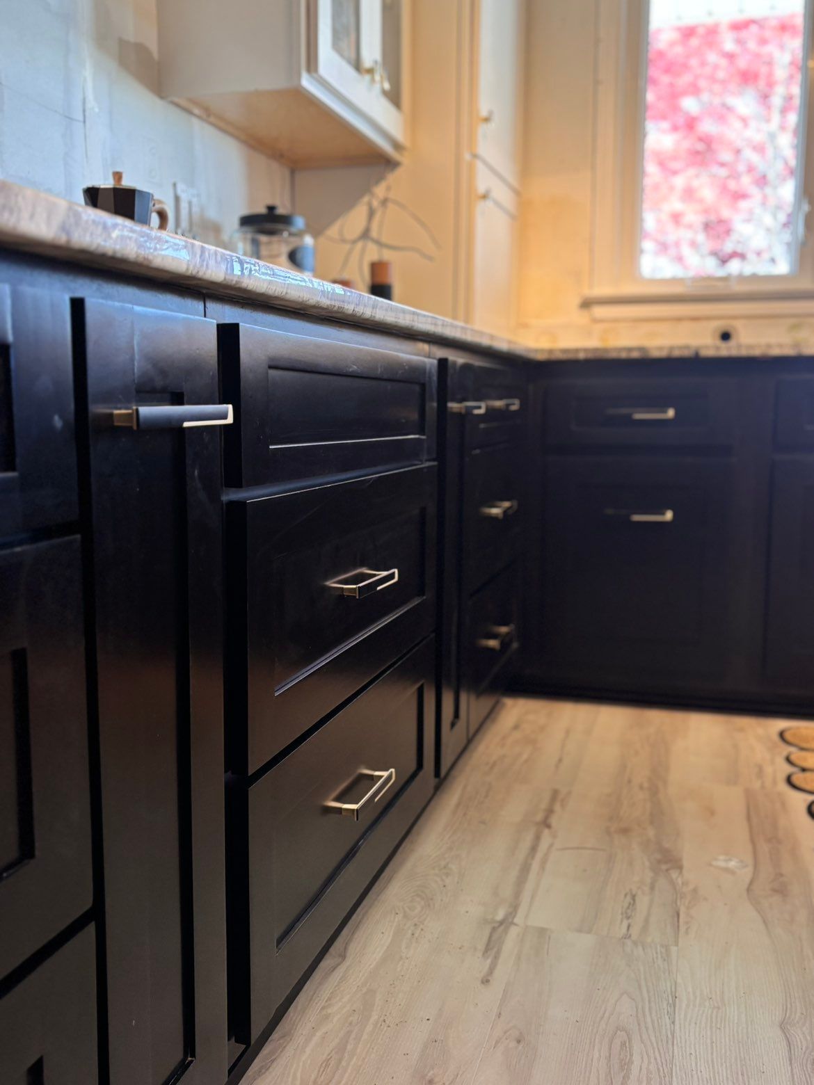 Modern kitchen with dark cabinets, light wood floor, and a sunlit window.