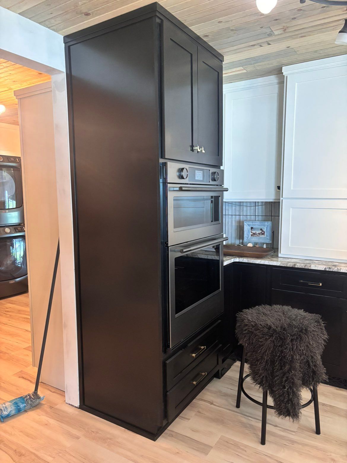 Tall black kitchen cabinet with built-in oven next to a white countertop and furry stool in a bright room