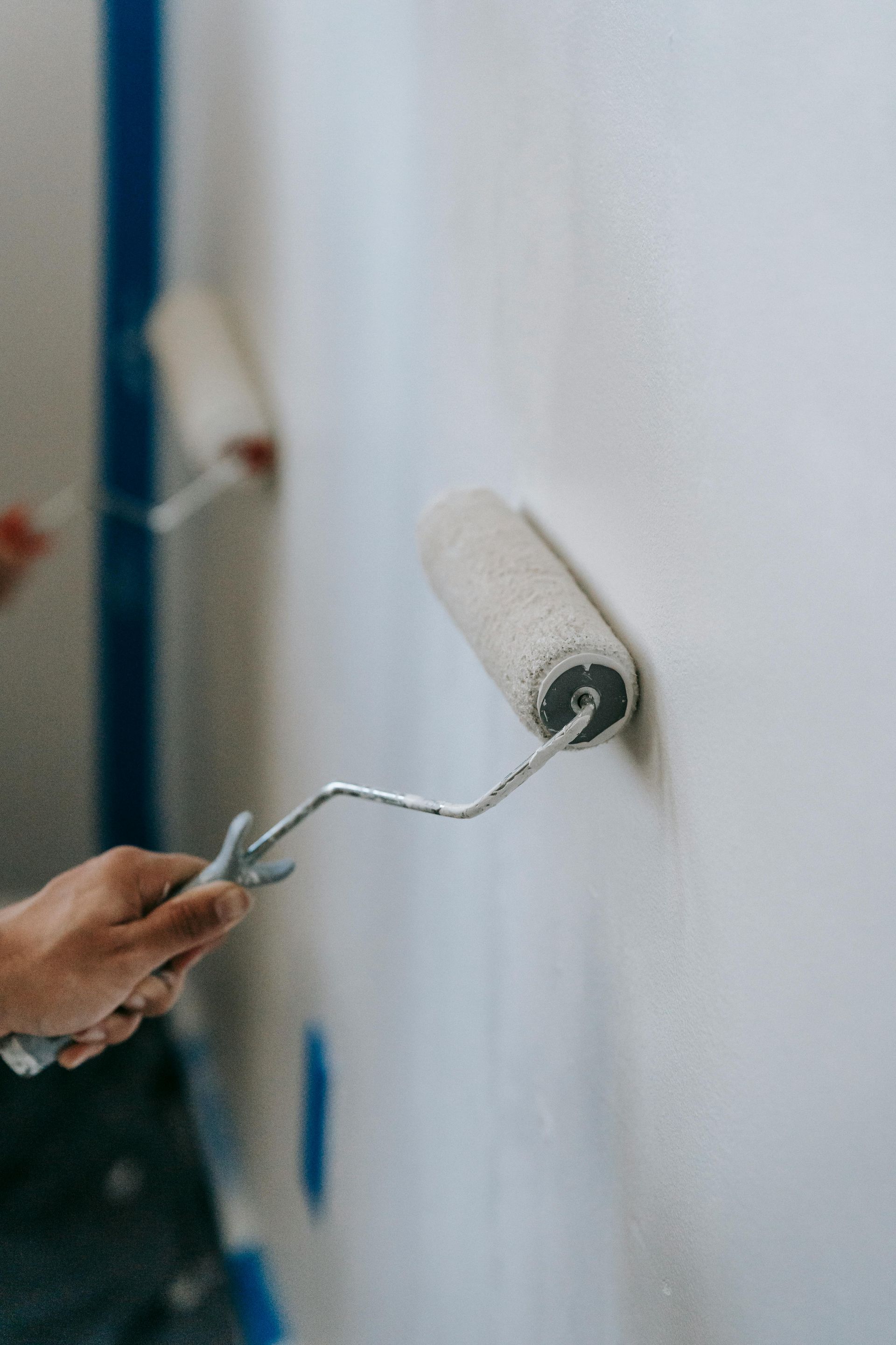 A hand rolls white paint on a wall with a paint roller, with blue painter’s tape along the edge.