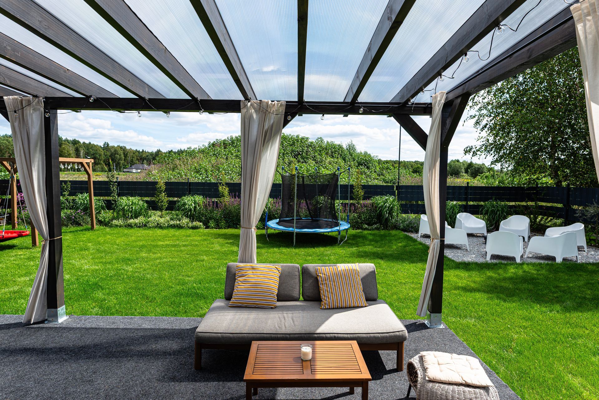 Covered patio with cushioned seating, striped canopy, green lawn, and trampoline in the backyard.