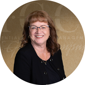 Woman with glasses smiles; wearing a black jacket and a necklace, against a brown backdrop with text.