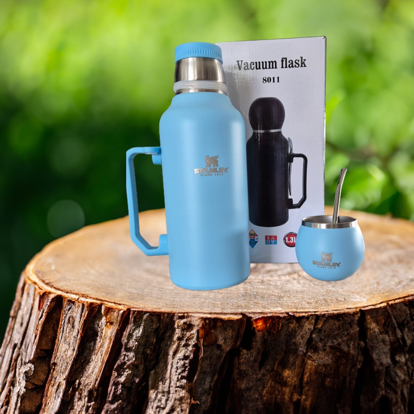 Un termo azul claro, un mate y una pajita de acero inoxidable se encuentran sobre un tocón de madera con un fondo verde.