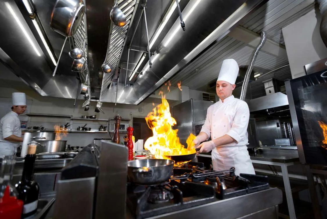 A chef is cooking in a kitchen with flames coming out of a pan.
