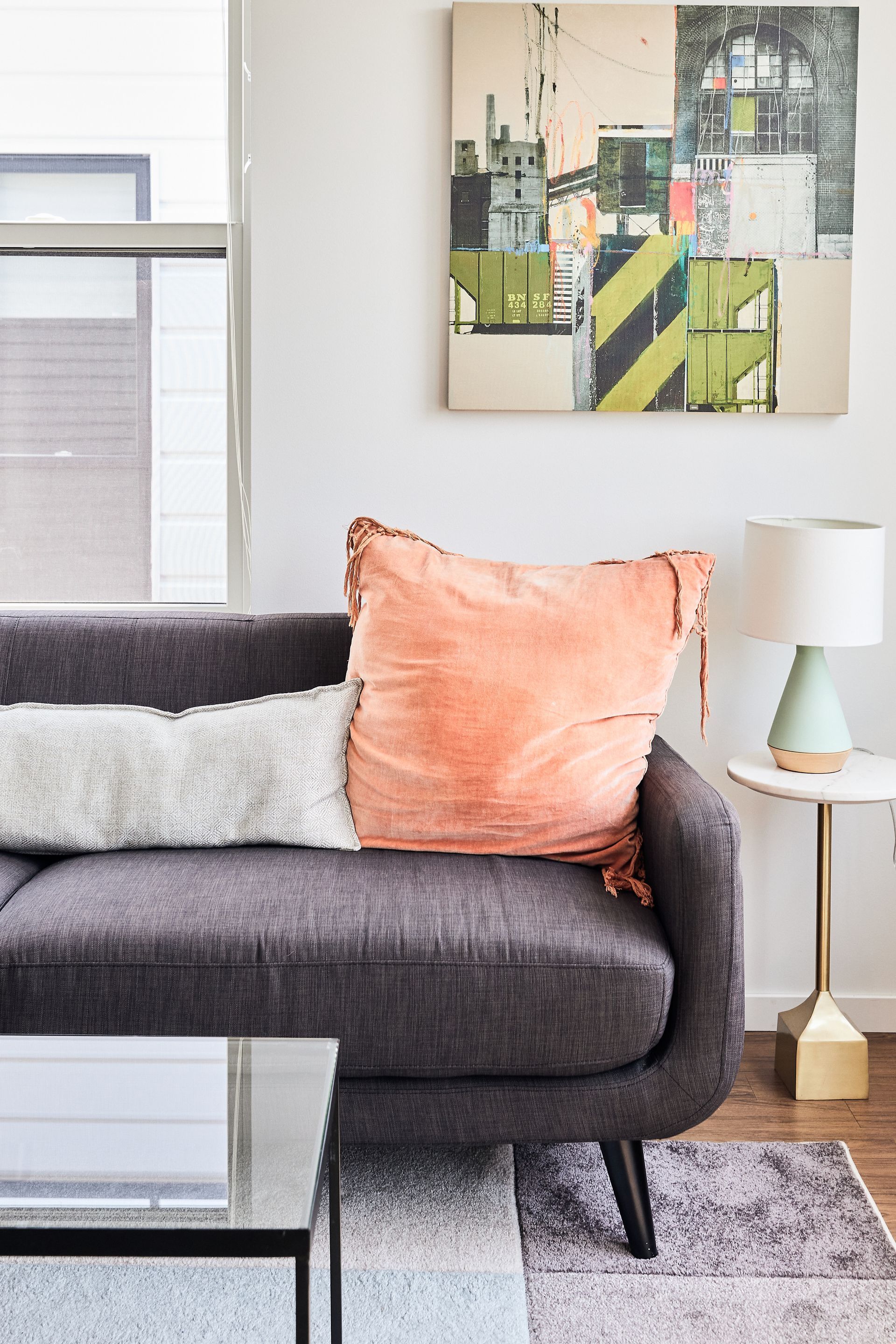 Gray couch with coral pillow, white pillow, painting, side table, and lamp.