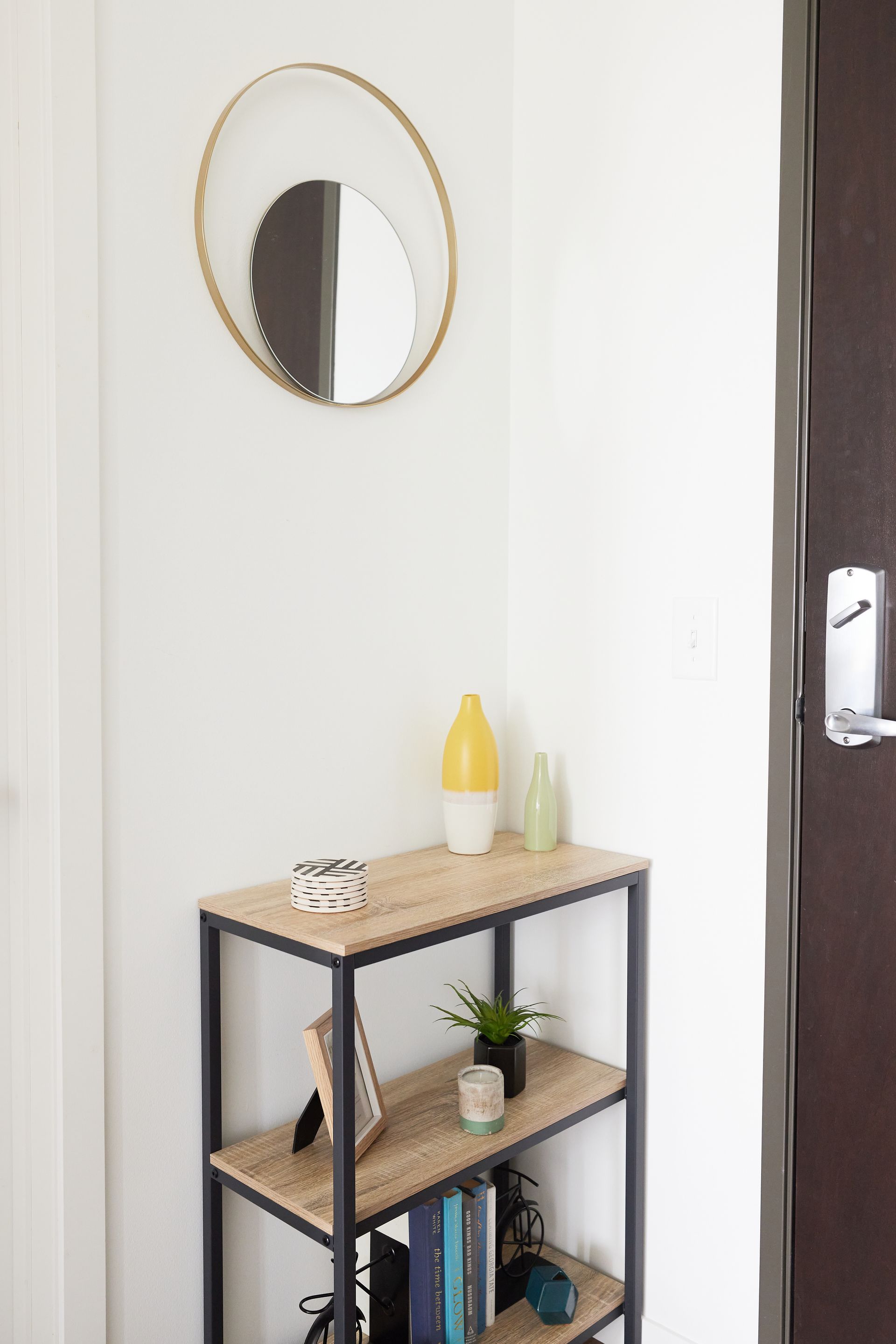 A small entryway with a shelf holding decor, a circular mirror, and a brown door.
