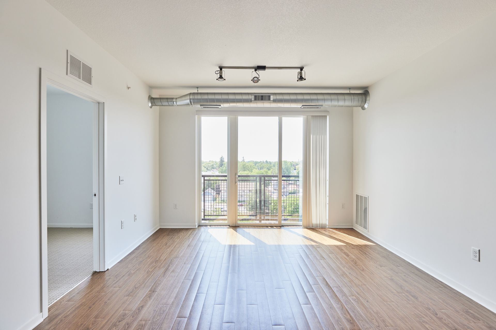 Empty living room with wood floors, large windows, and a balcony.