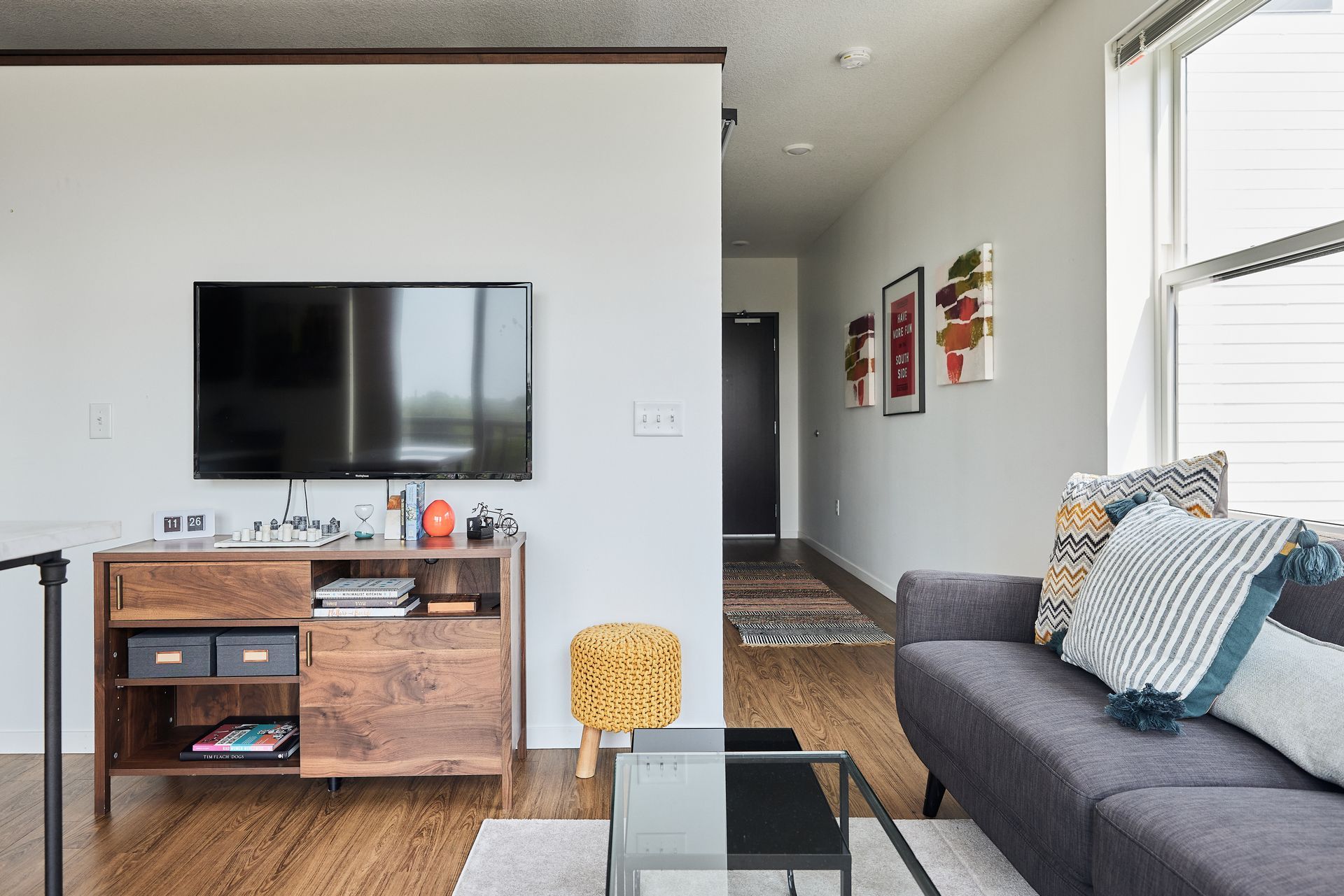 Living room with TV, wooden cabinet, gray couch, and hallway with artwork.