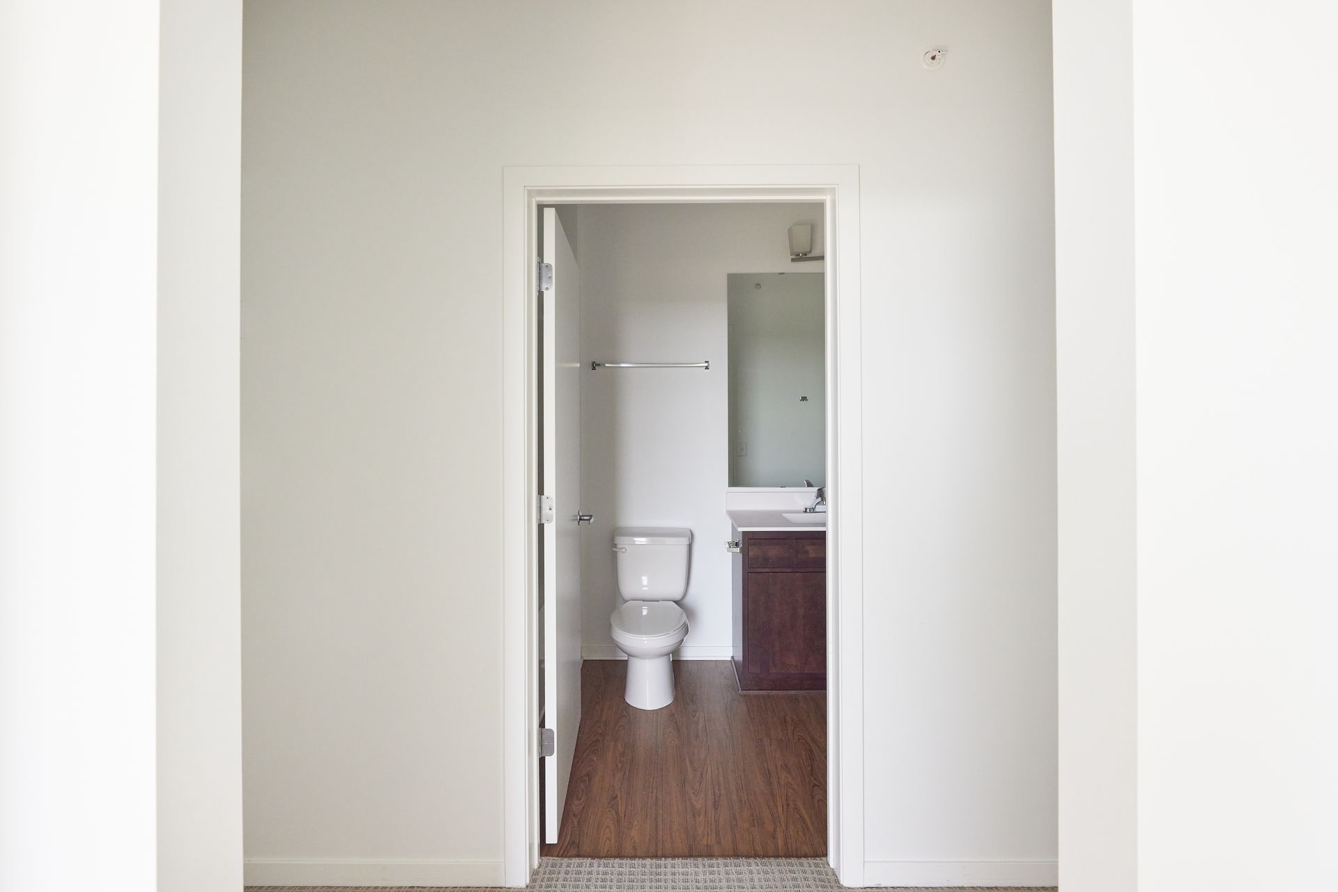 Open doorway to a bathroom with a toilet, mirror, and vanity; hardwood floors.