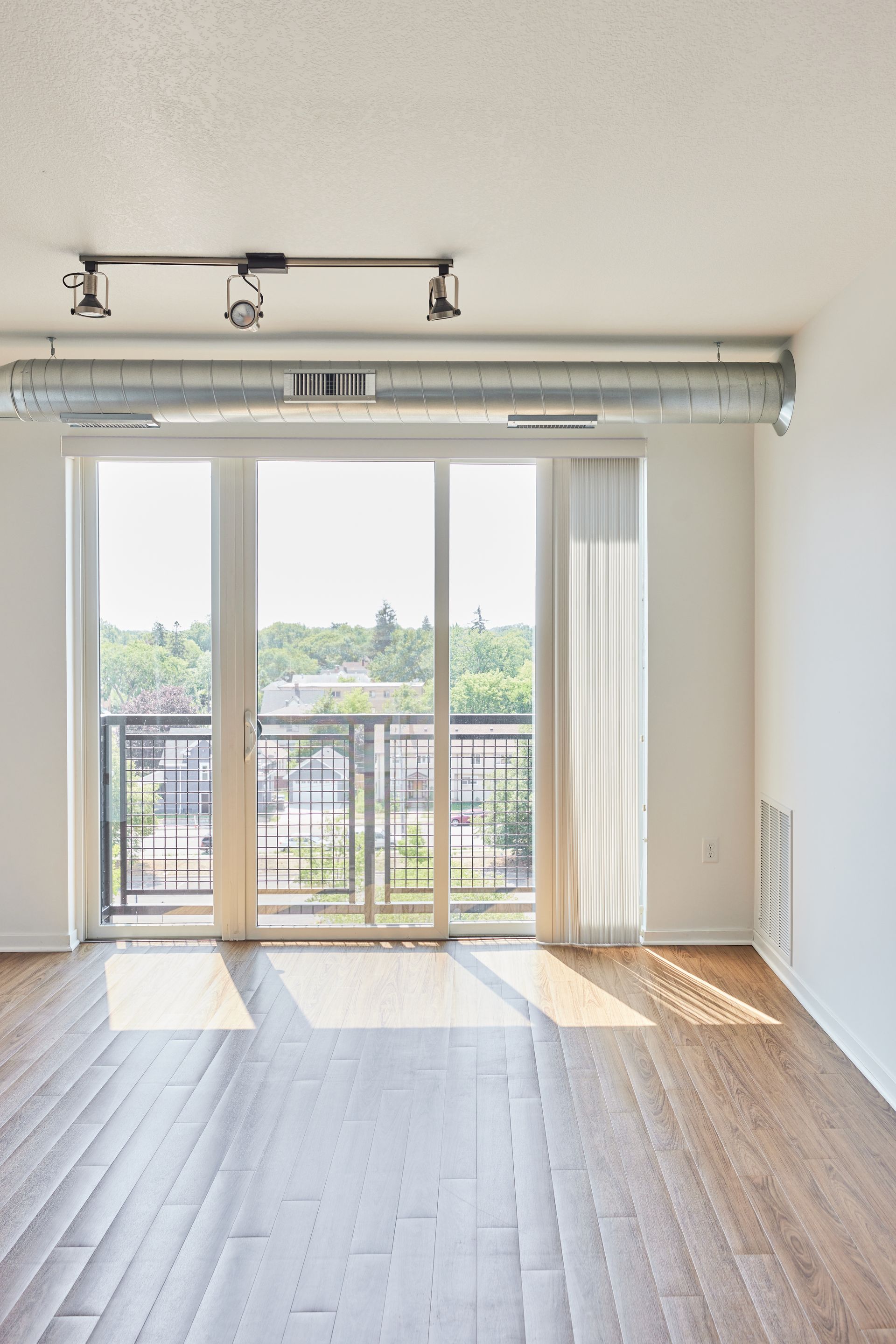 Empty sunlit room with balcony access, hardwood floors, and exposed ductwork.