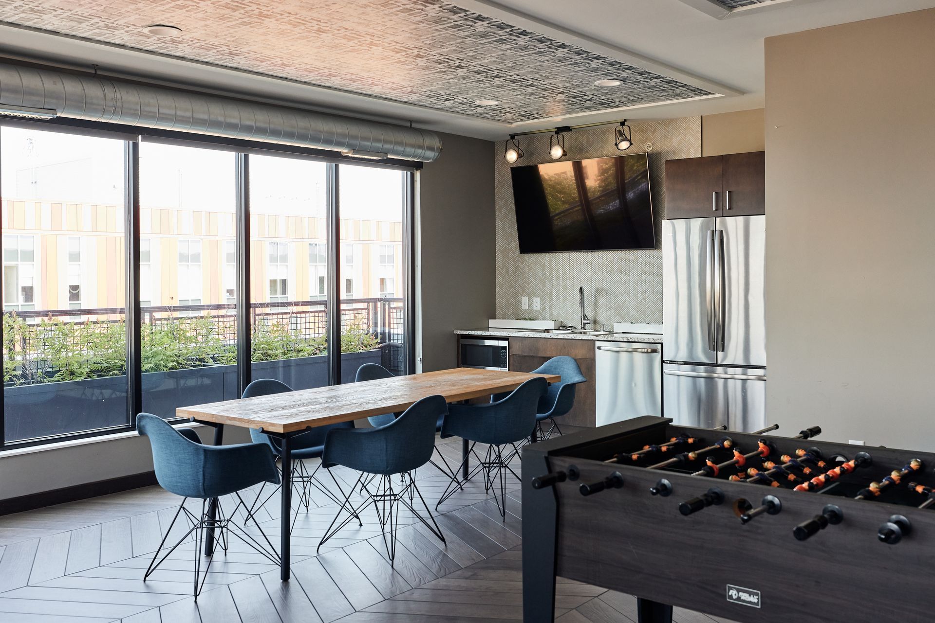 A modern lounge with a foosball table, dining table, kitchen, and a large window with a cityscape view.