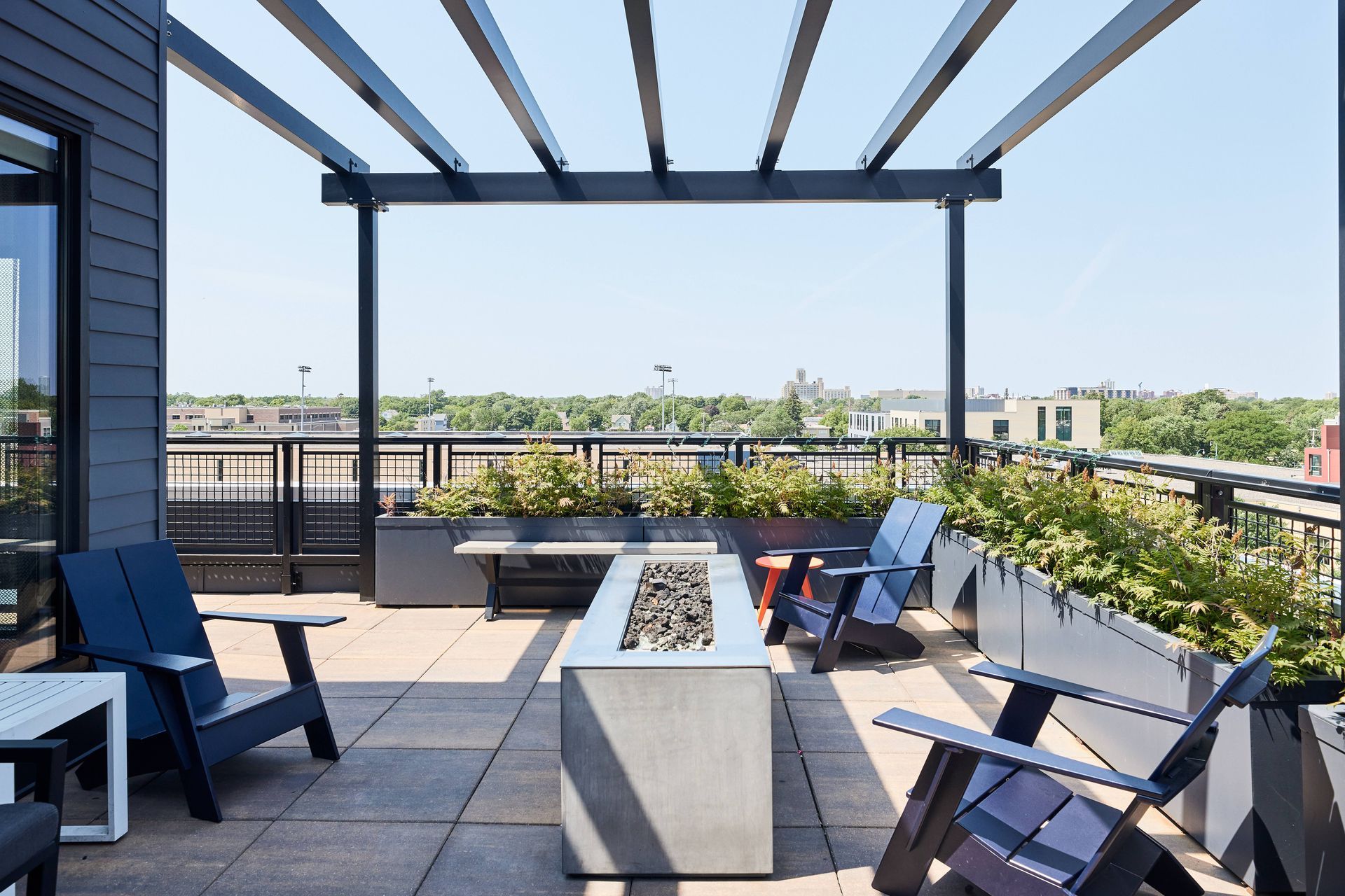 Rooftop patio with modern furniture, fire pit, and pergola overlooking cityscape on a sunny day.