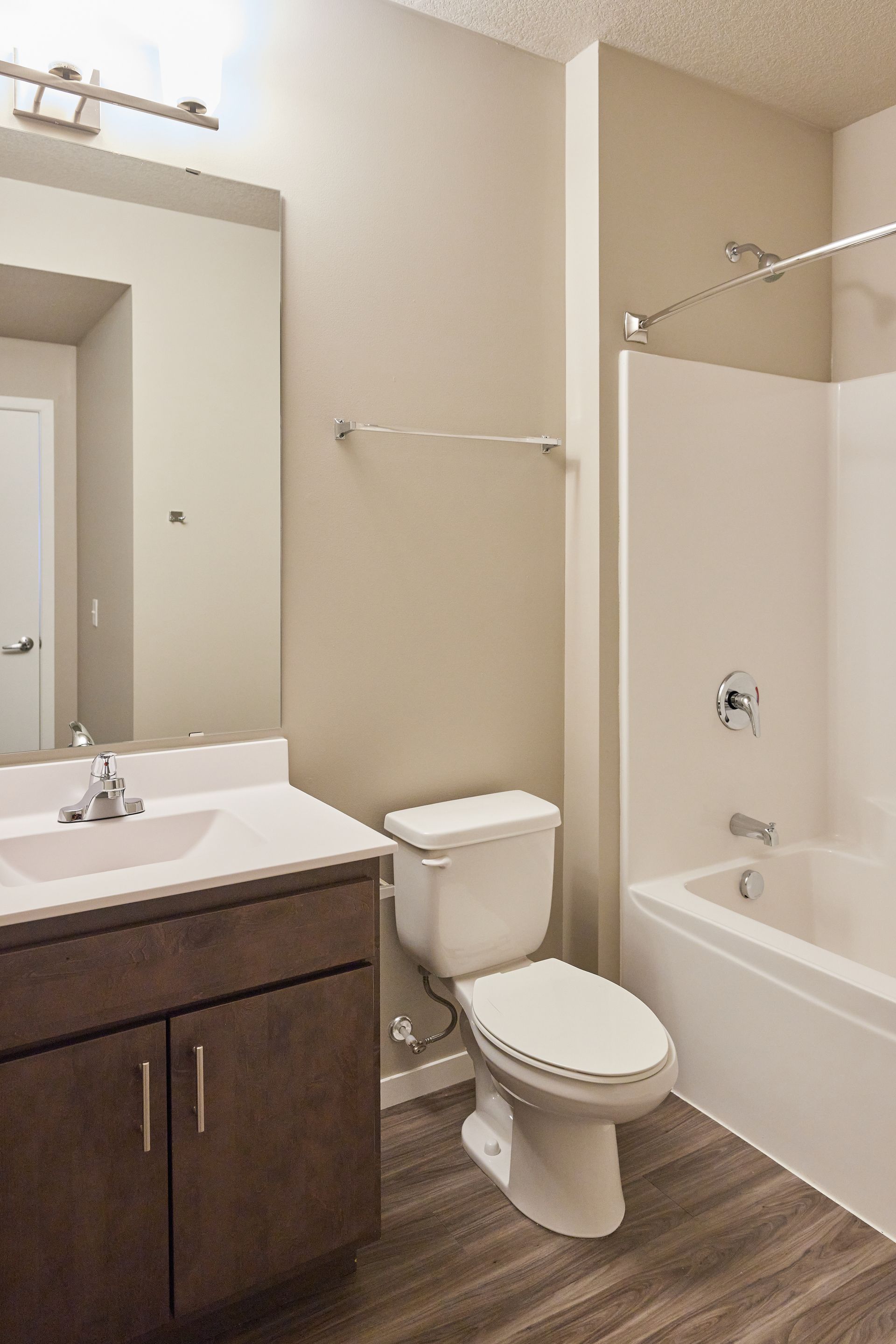 Bathroom with white tub, toilet, and sink. Brown vanity and light gray walls, with wooden floor.