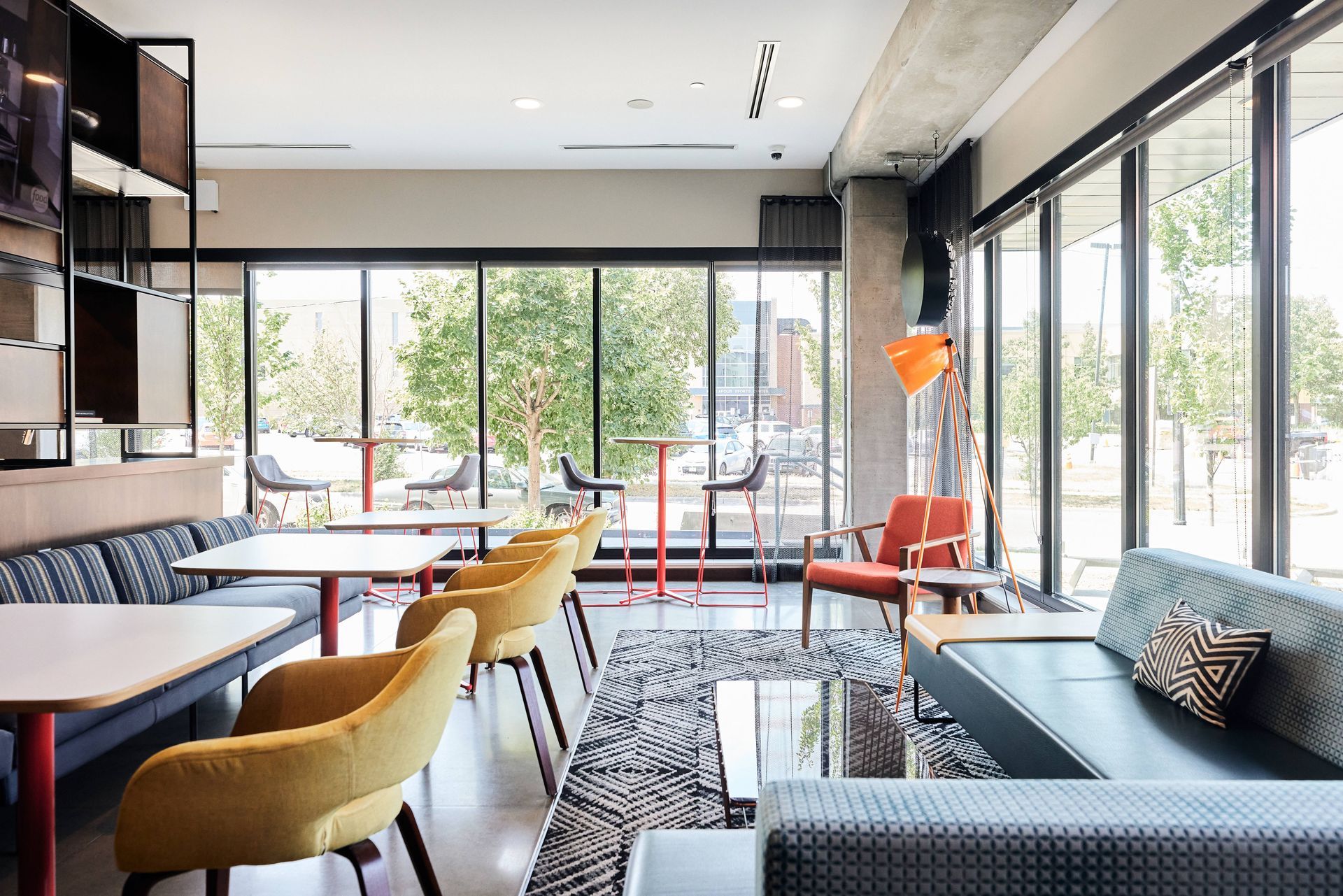 Modern cafe interior with floor-to-ceiling windows, seating in various colors and styles, and a view of outside trees.