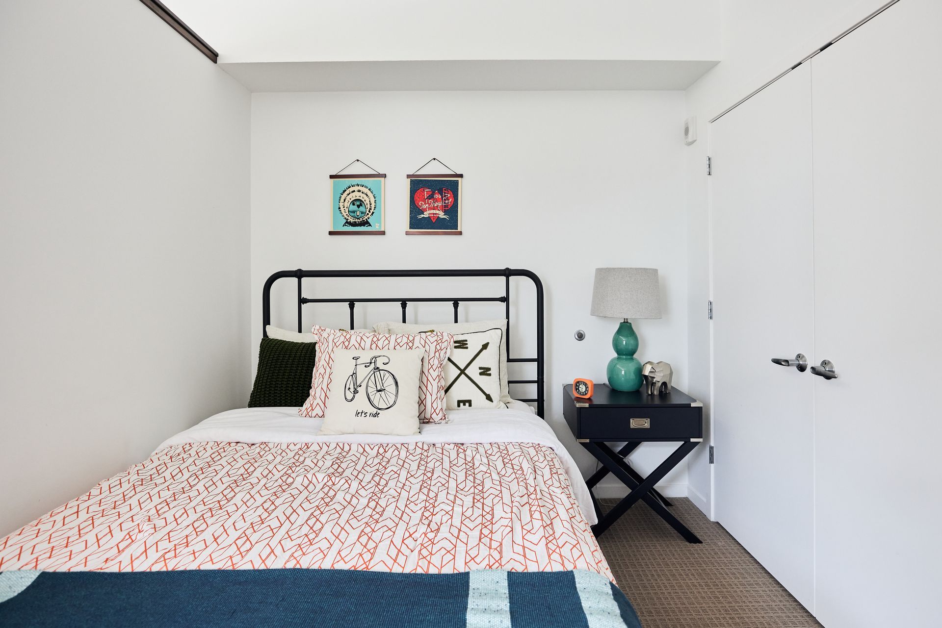 Bedroom with metal bed frame, patterned bedding, artwork, and a teal lamp on a nightstand.