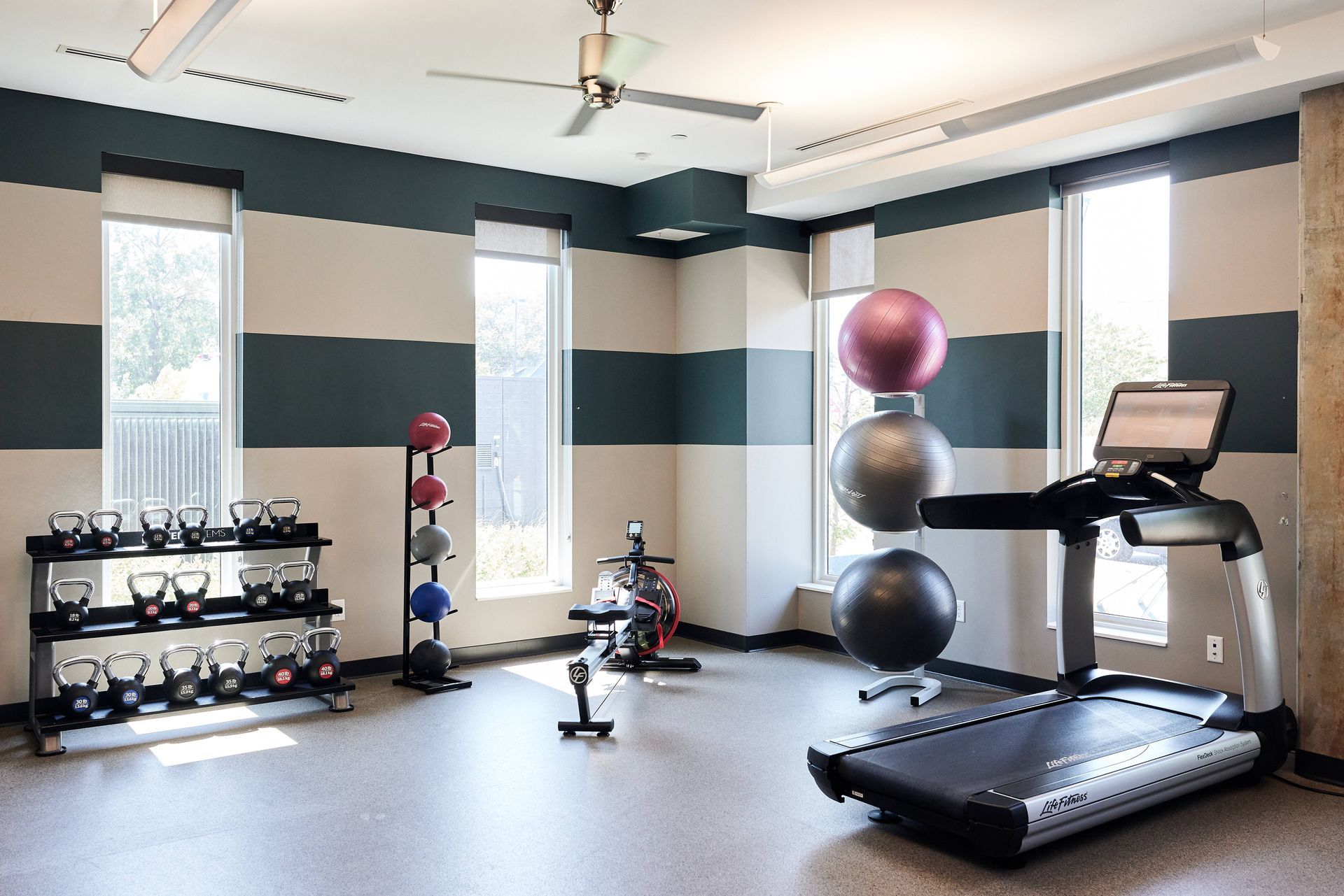 Gym with fitness equipment: kettlebells, exercise balls, treadmill, bike, windows, and striped wall.