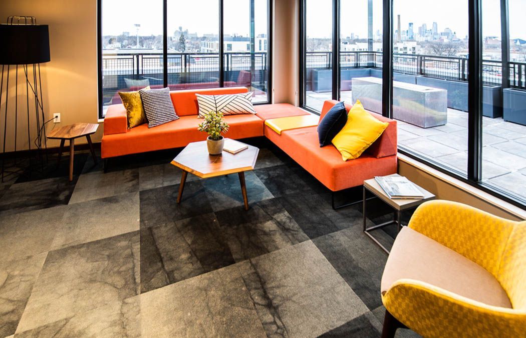 Modern lounge with orange sectional, yellow chair, patterned gray floors, large windows, and city view.