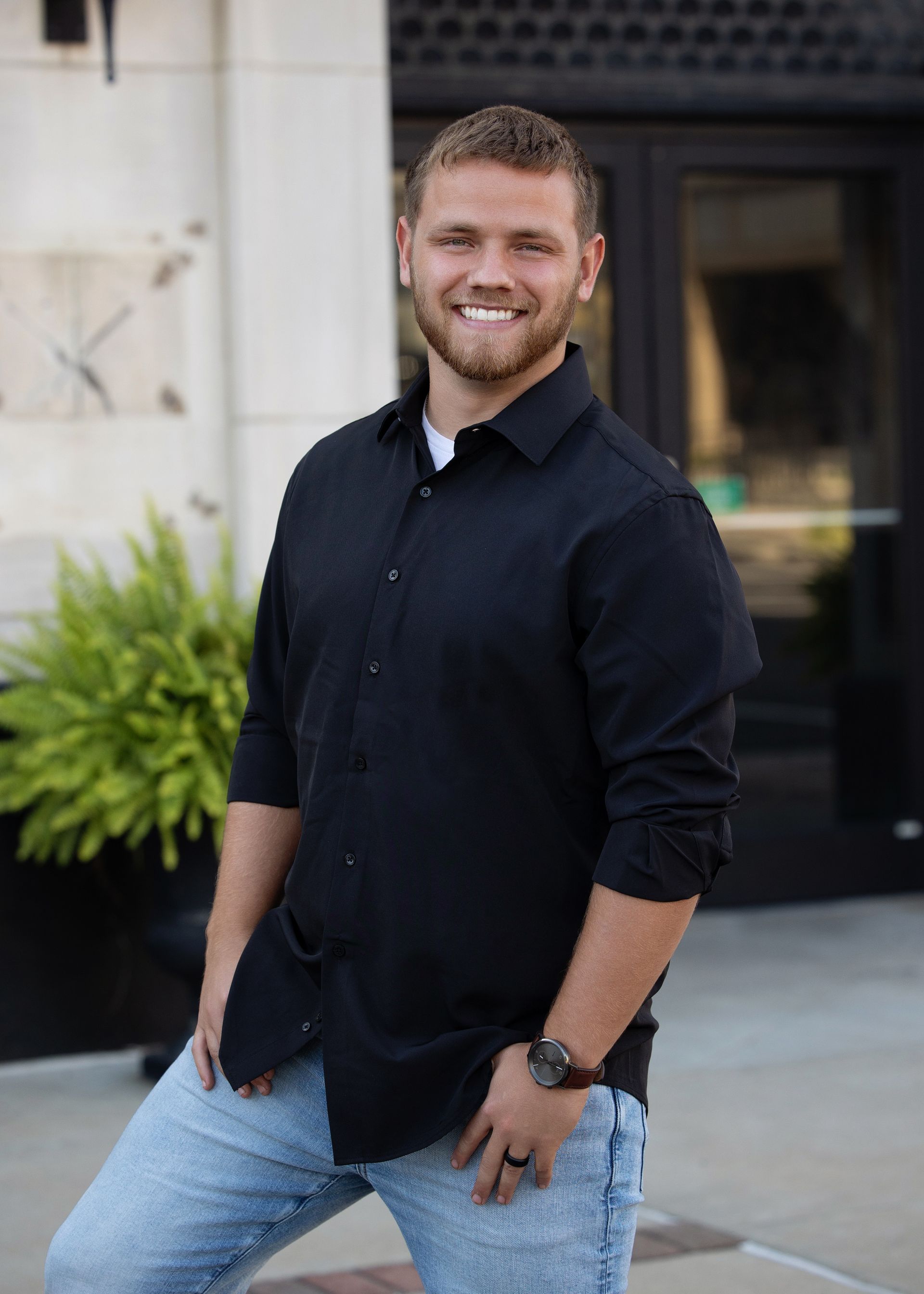 A man in a black shirt and blue jeans is standing in front of a building.
