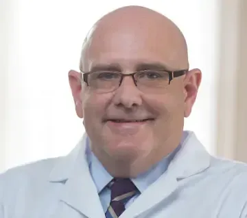 A bald man wearing glasses and a white coat and tie is smiling.