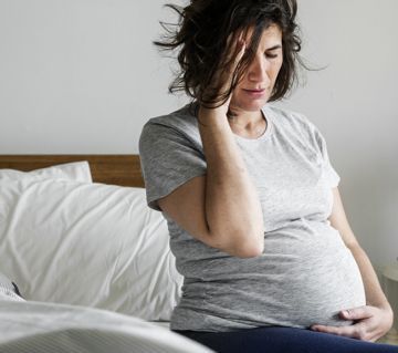 A pregnant woman is sitting on a bed holding her head.