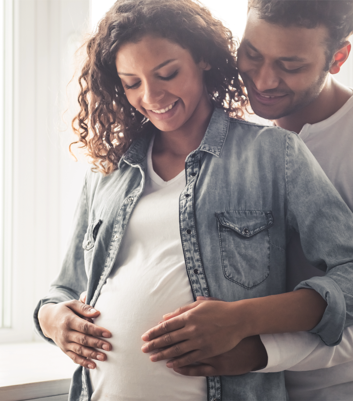 A man is hugging a pregnant woman in front of a window