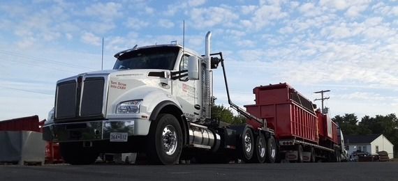 A white semi-truck with a hook lift system pulling a flatbed trailer loaded with red dumpsters against a cloudy sky.