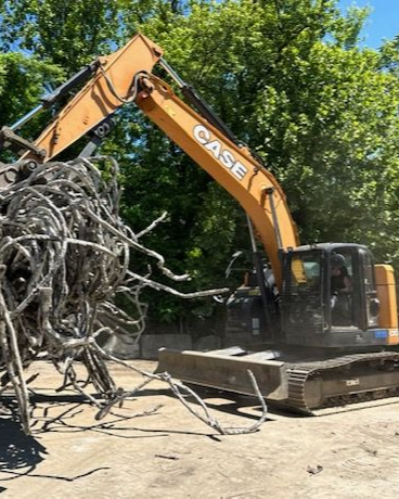 A yellow Case excavator lifts a large, tangled metal structure against a backdrop of green trees.