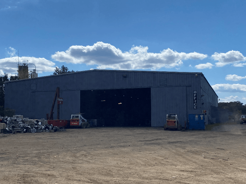 A large grey industrial warehouse with an open entrance, two small loaders parked in front, under a blue sky with clouds.