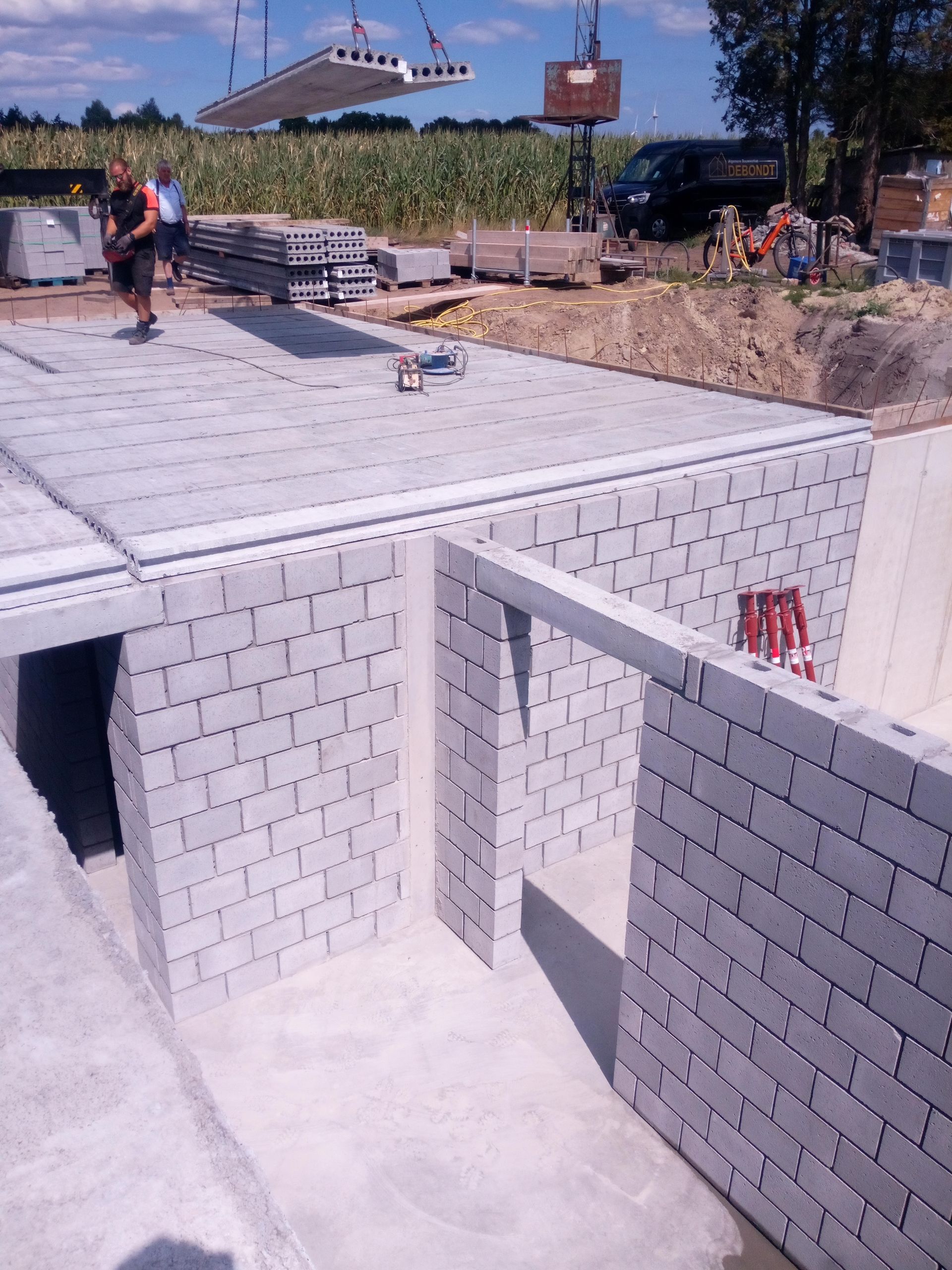 Een wit bakstenen betonnen constructie met een open deuropening op een bouwterrein; op de achtergrond zijn bouwvakkers zichtbaar.