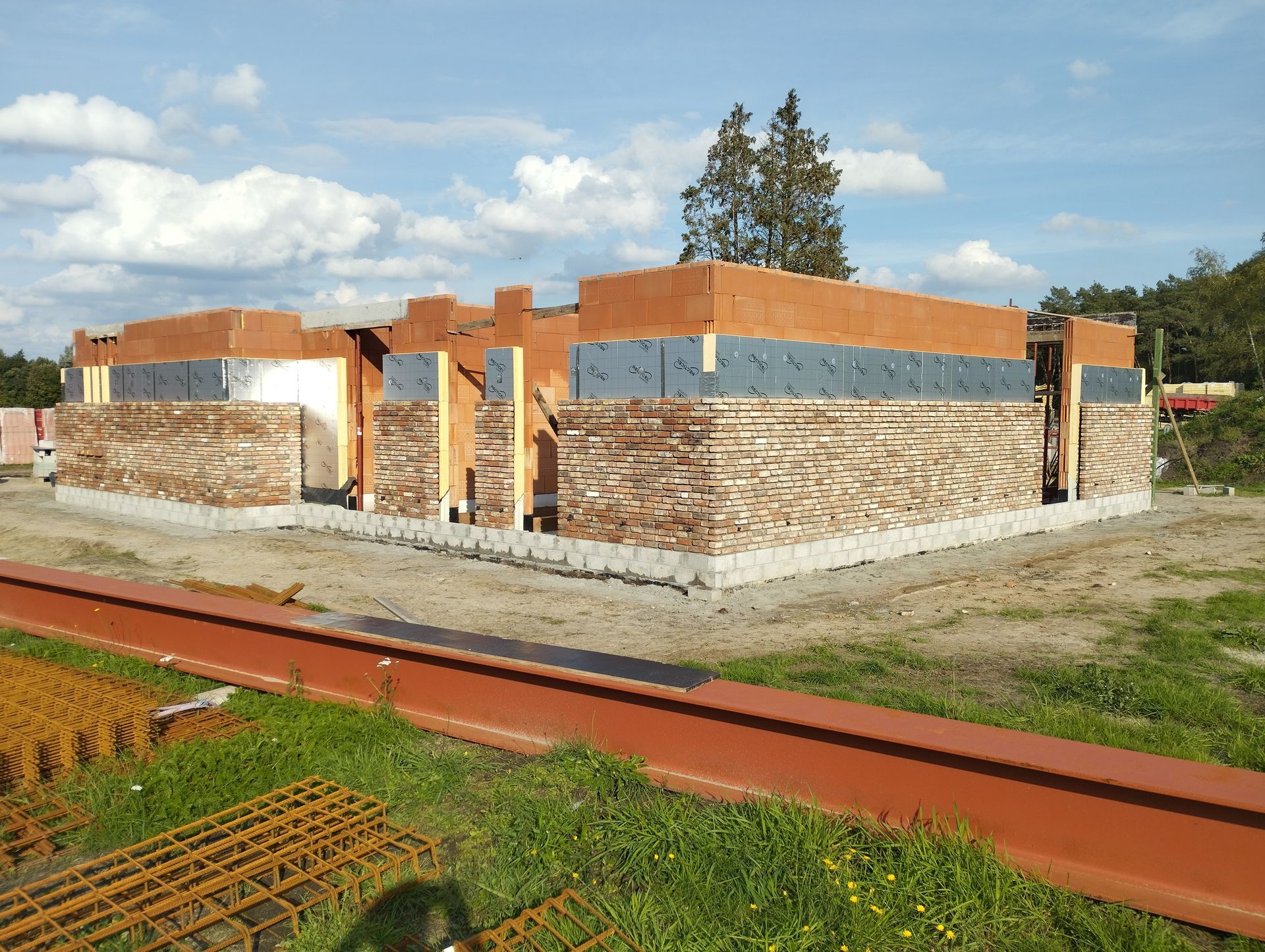 Een gedeeltelijk gebouwd bakstenen huis met betonnen fundering onder een blauwe hemel, gezien vanaf een onverharde weg.