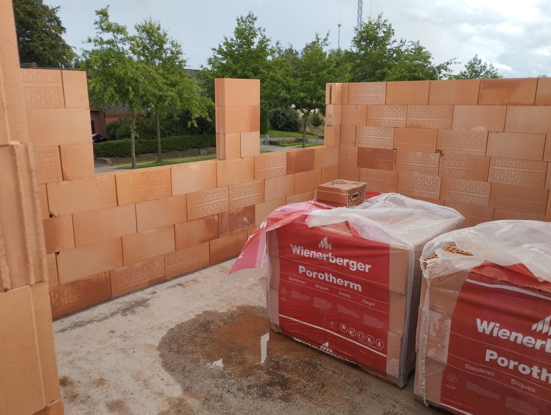 Stapels oranje bouwstenen bij een gedeeltelijk gebouwde muur, met op de voorgrond ingepakt palletmateriaal.