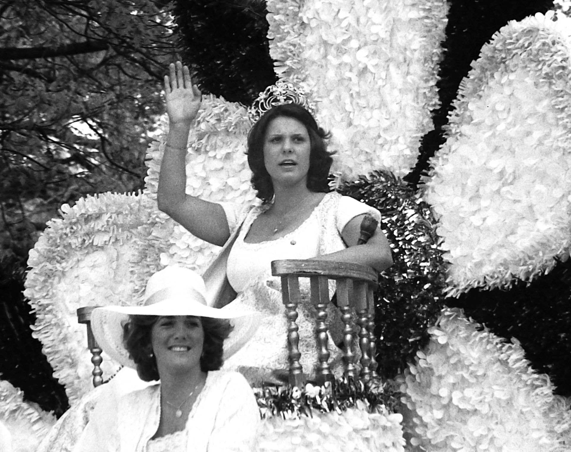 Two women wave from a flower-decorated float. One raises her arm. The other wears a hat and smiles.
