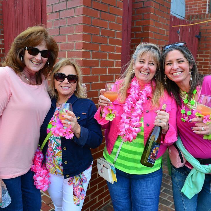 Four women smiling, holding drinks, wearing colorful leis and sunglasses near a brick wall.