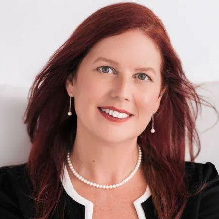 Woman with auburn hair, wearing a pearl necklace, smiling, and looking at the camera.