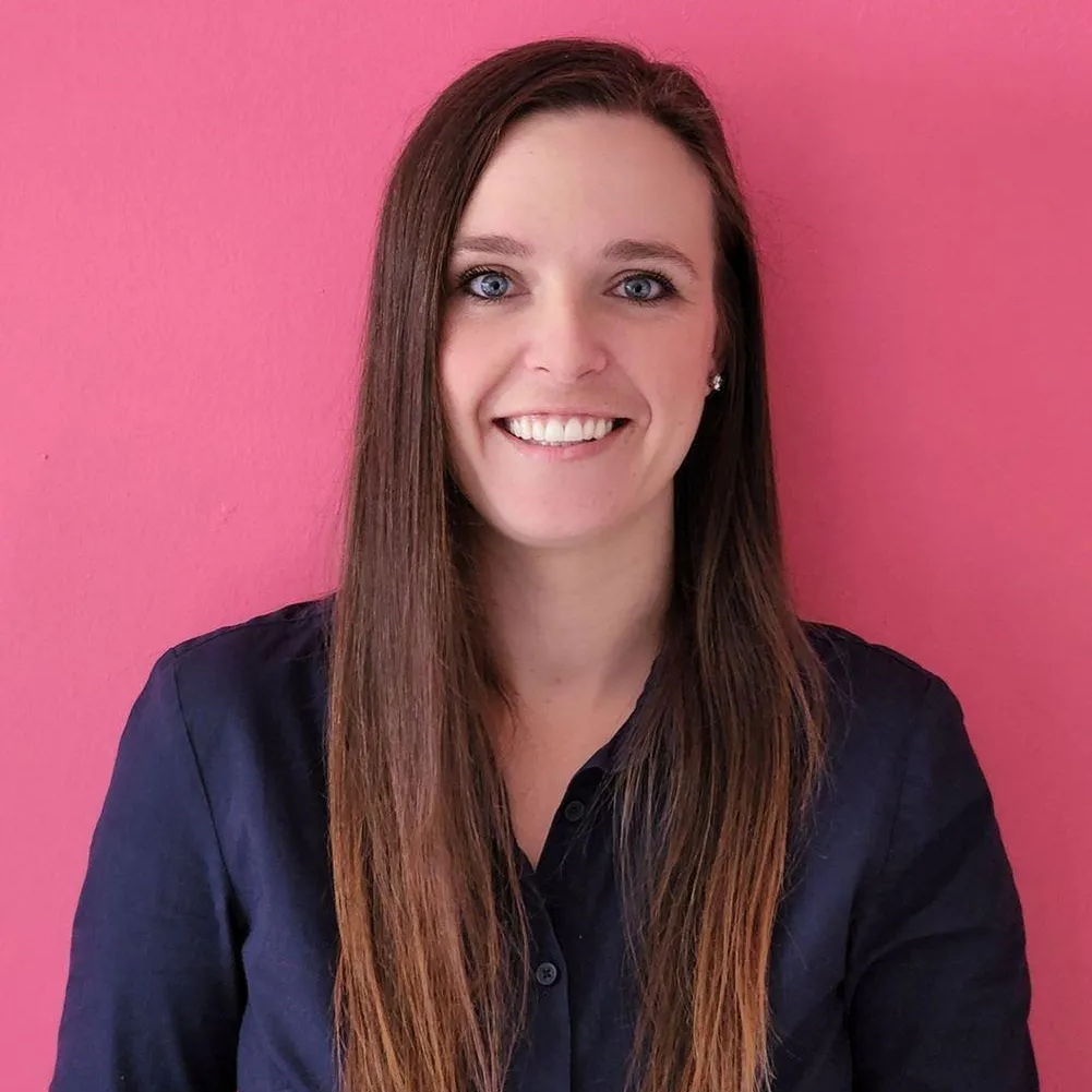Woman with long brown hair smiling in front of a pink wall, wearing a navy blue shirt.