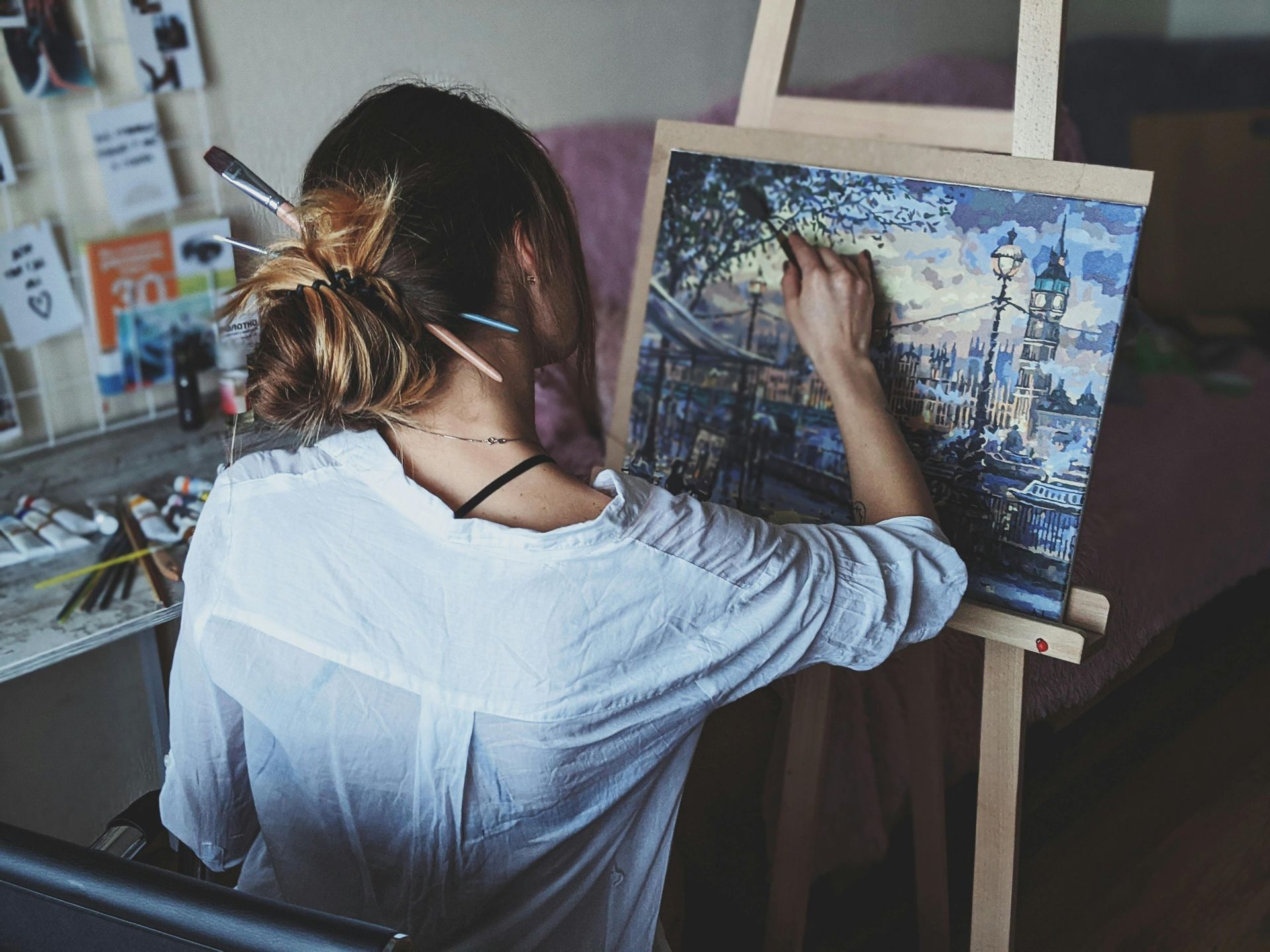 Woman paints at easel, brushes in her hair. She wears a white shirt and works on a landscape.