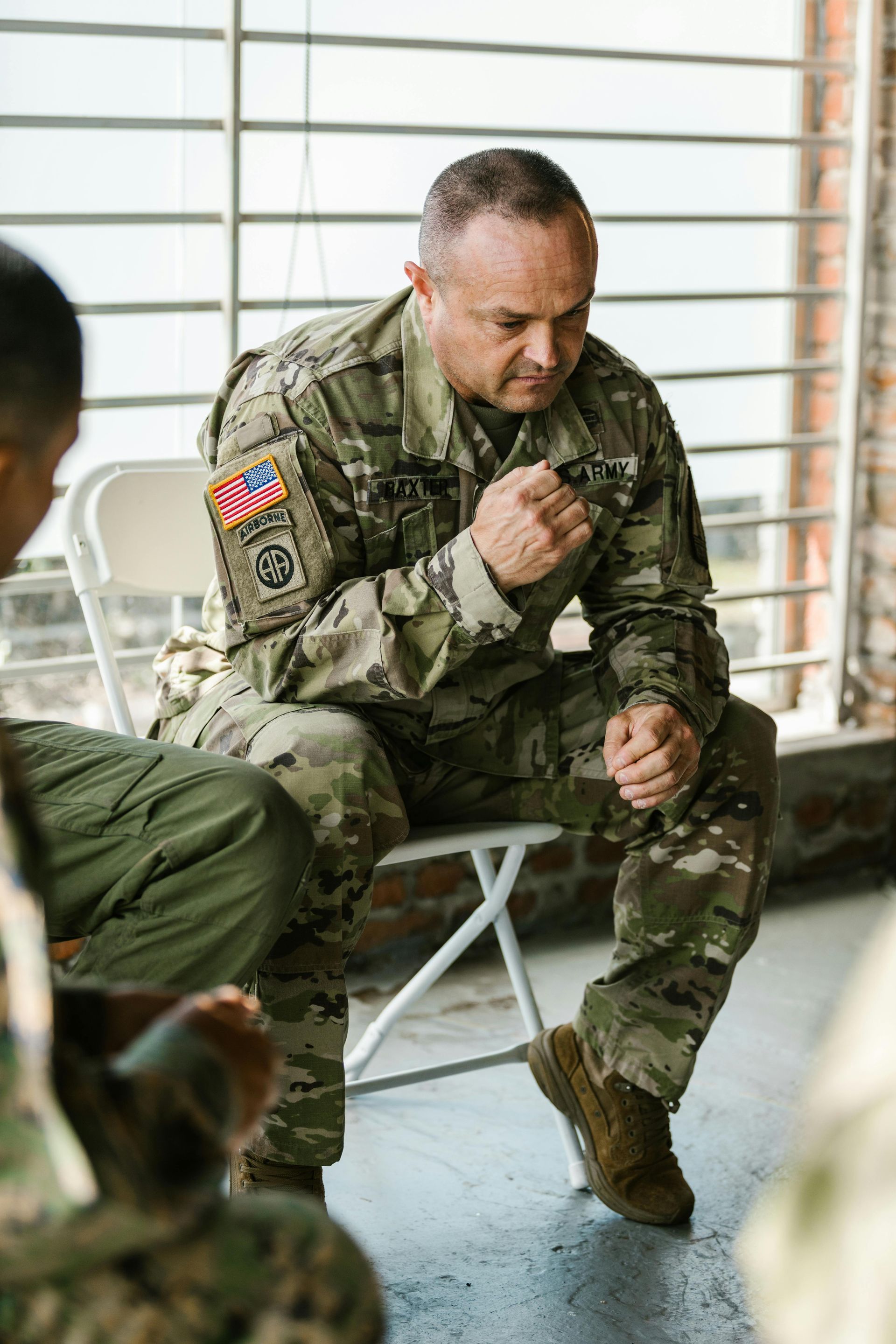 Soldier in camouflage uniform seated, looking down, hands clasped, in discussion with others outdoors.