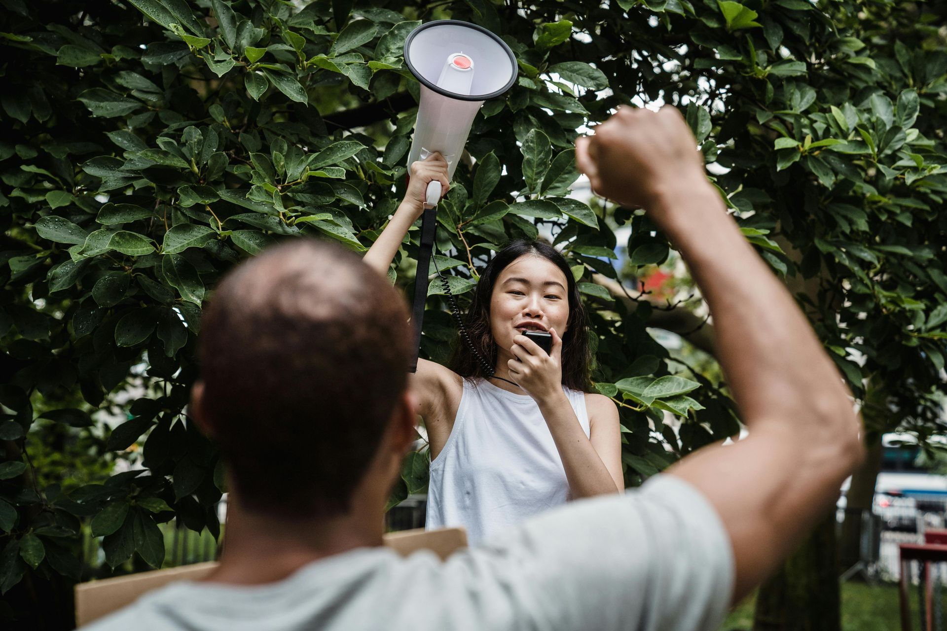 Woman speaking into megaphone, arm raised. Man in foreground raises fist. Green trees in background.