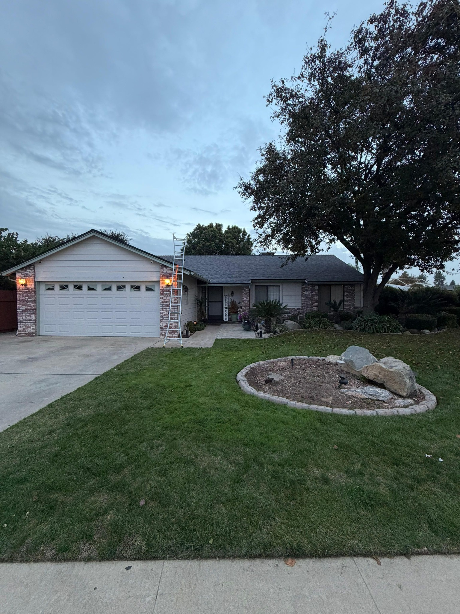 Single-story suburban house with a white garage, gray shingle roof, green lawn, and a large tree during twilight.