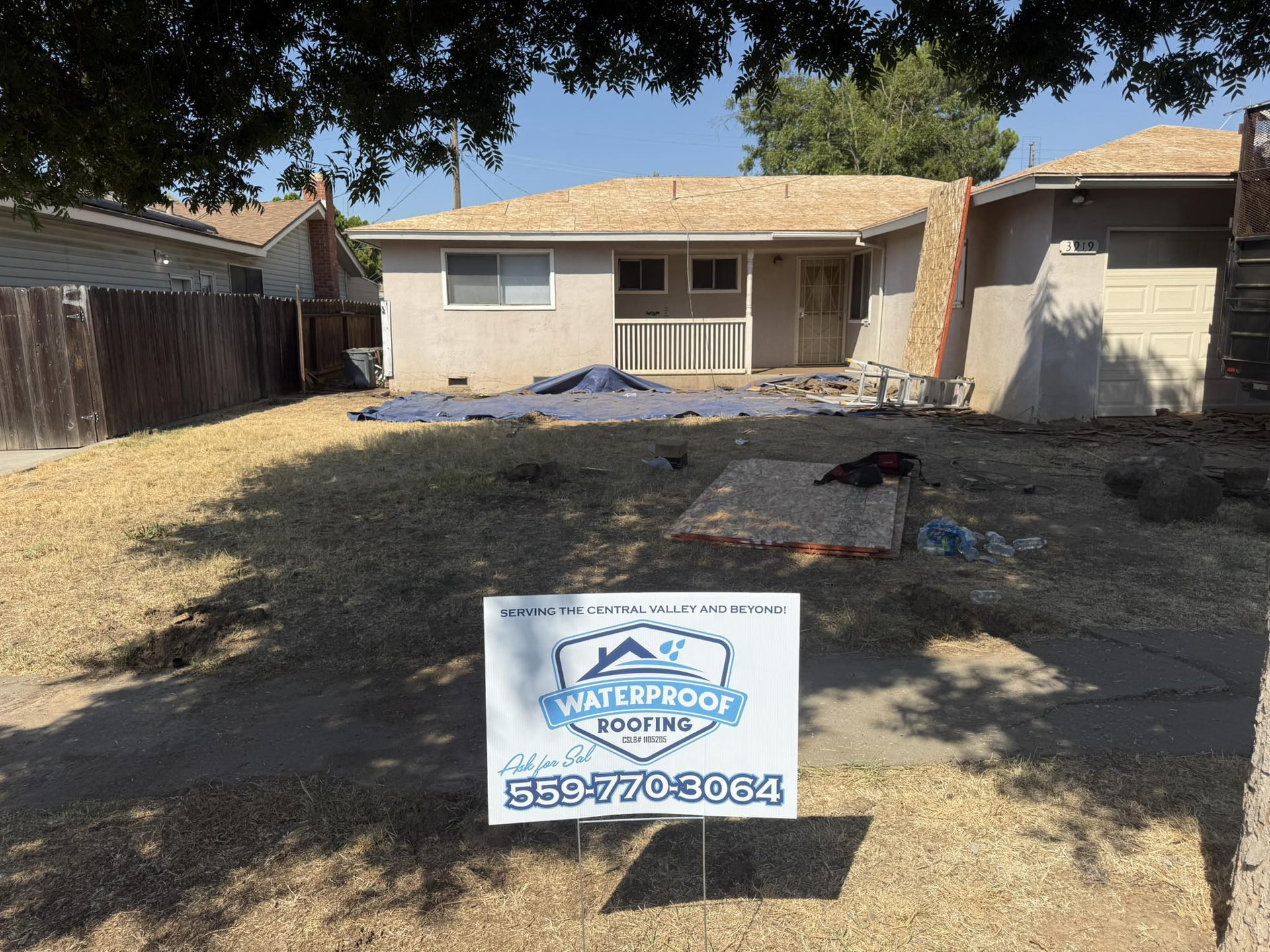 A beige house with a roof under renovation, seen from a lawn with a Waterproof Roofing sign in the foreground.