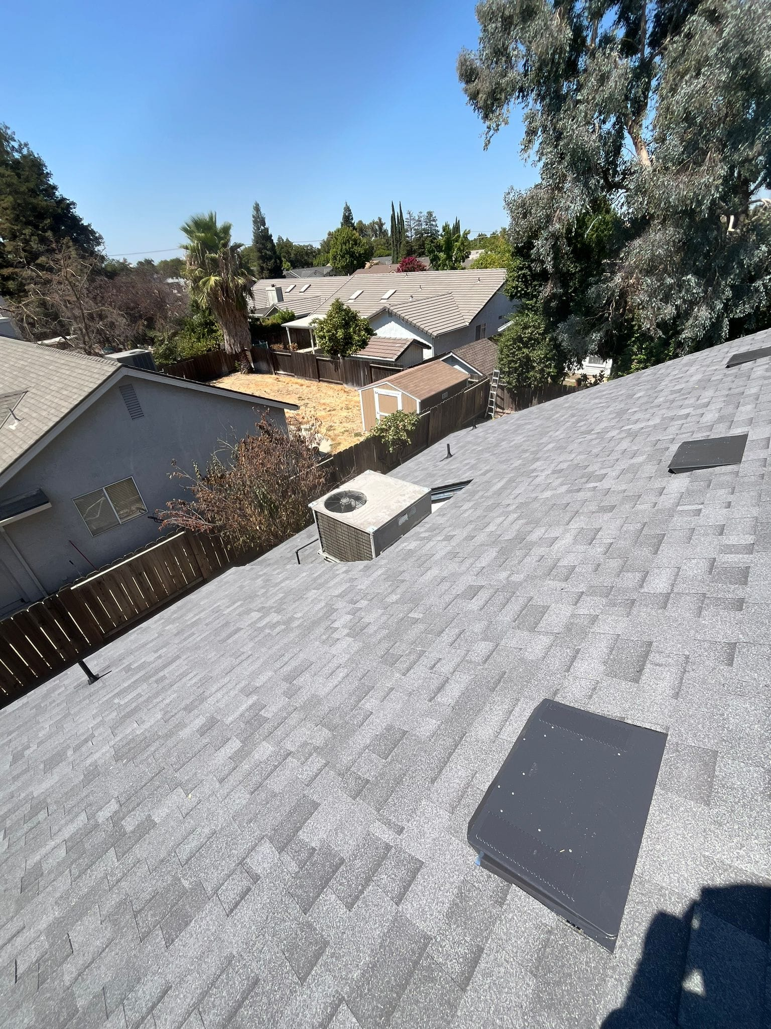 View of a gray shingled roof with a chimney and vents, other houses and trees in the background under a blue sky.