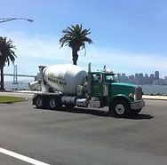 Green cement mixer truck parked on a sunny road by palm trees with a bridge and city skyline in the background Green cement mixer truck parked on a sunny road by palm trees with a bridge and city skyline in the background