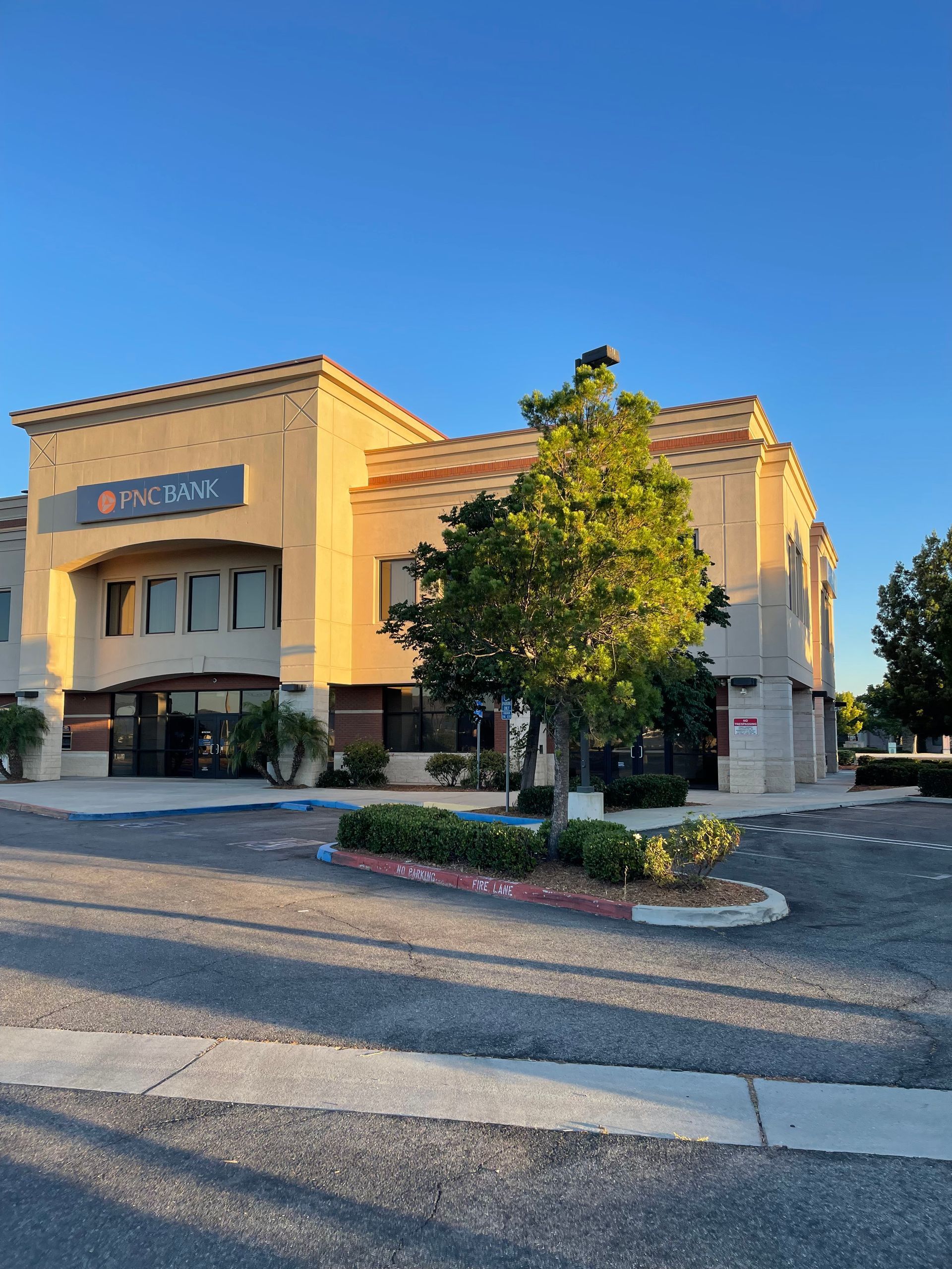 Two-story tan office building under a clear blue sky. A tree and bushes are in front of the building.