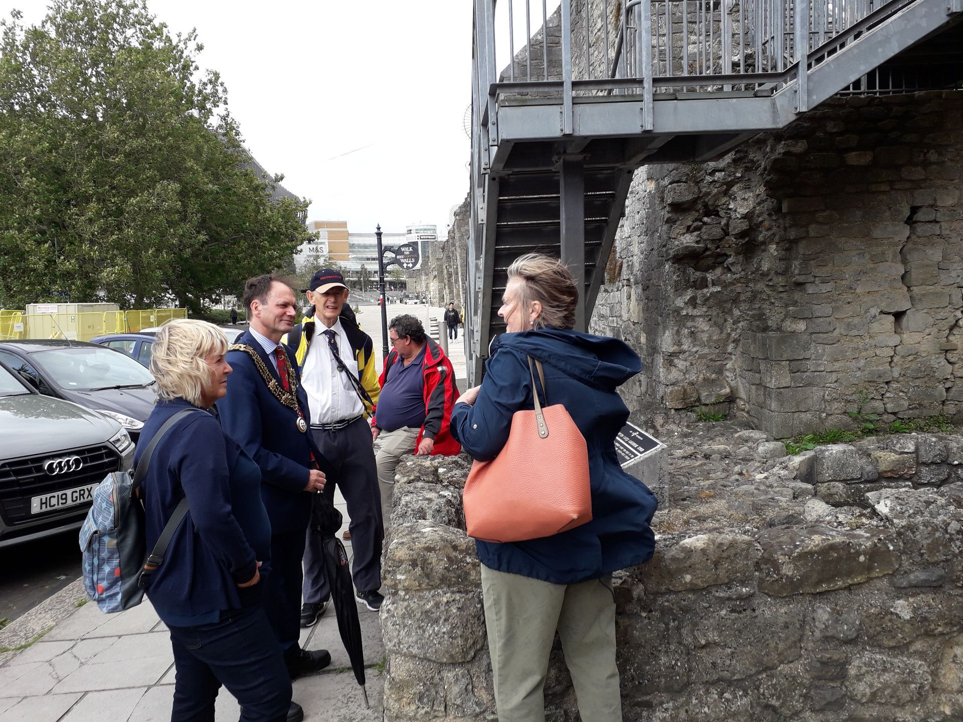 A group of people are standing on a sidewalk next to a stone wall.