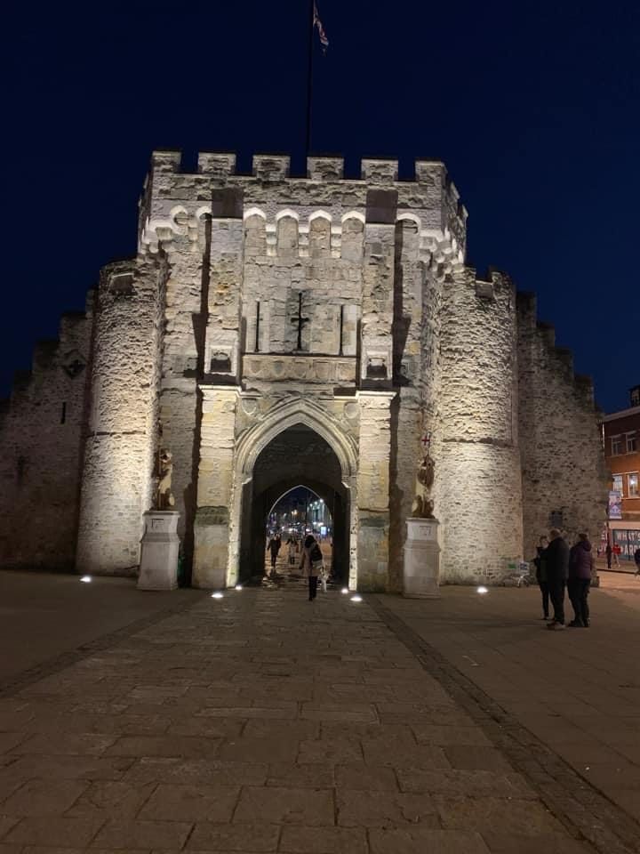 A castle gate is lit up at night with people standing in front of it.