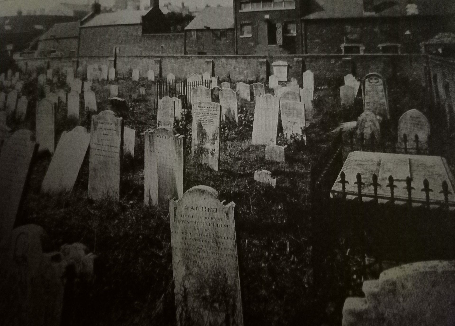 A black and white photo of a cemetery with houses in the background.