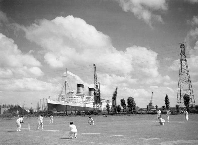 A black and white photo of people playing cricket with a large ship in the background