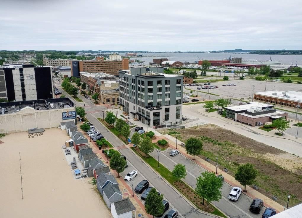 Aerial view of a coastal city street with buildings, parked cars, trees, and a waterfront in the distance