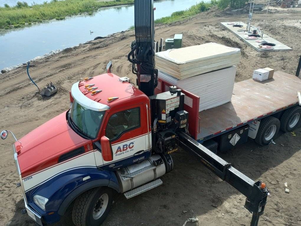 Red and blue truck with crane lifting stacked white panels beside a river.