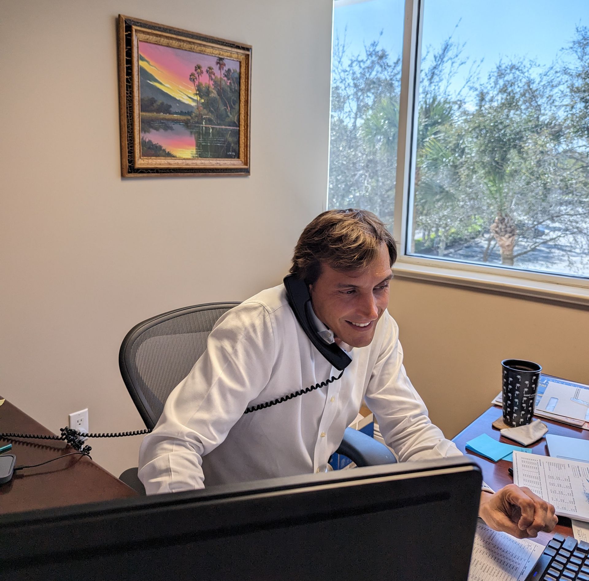 Man in a suit smiling, with red tie and white shirt. Standing indoors near a green plant.